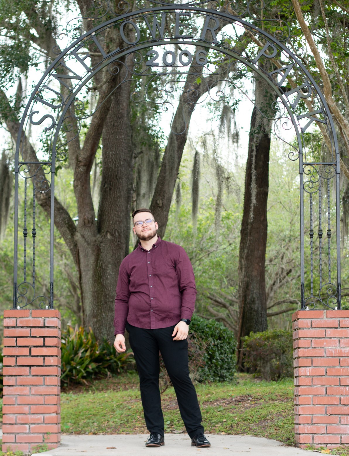 Profile image of Wade Hart, full body, standing under an entrance outside made of a brick wall and a wire arch. Trees are in the background.