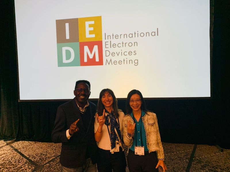 Professor Jean Anne Incorvia standing in front of a screen reading “IEDM” with Xiuling Li and Deji Akinwande on the sides of her, throwing “hook’em horns” hand gestures.